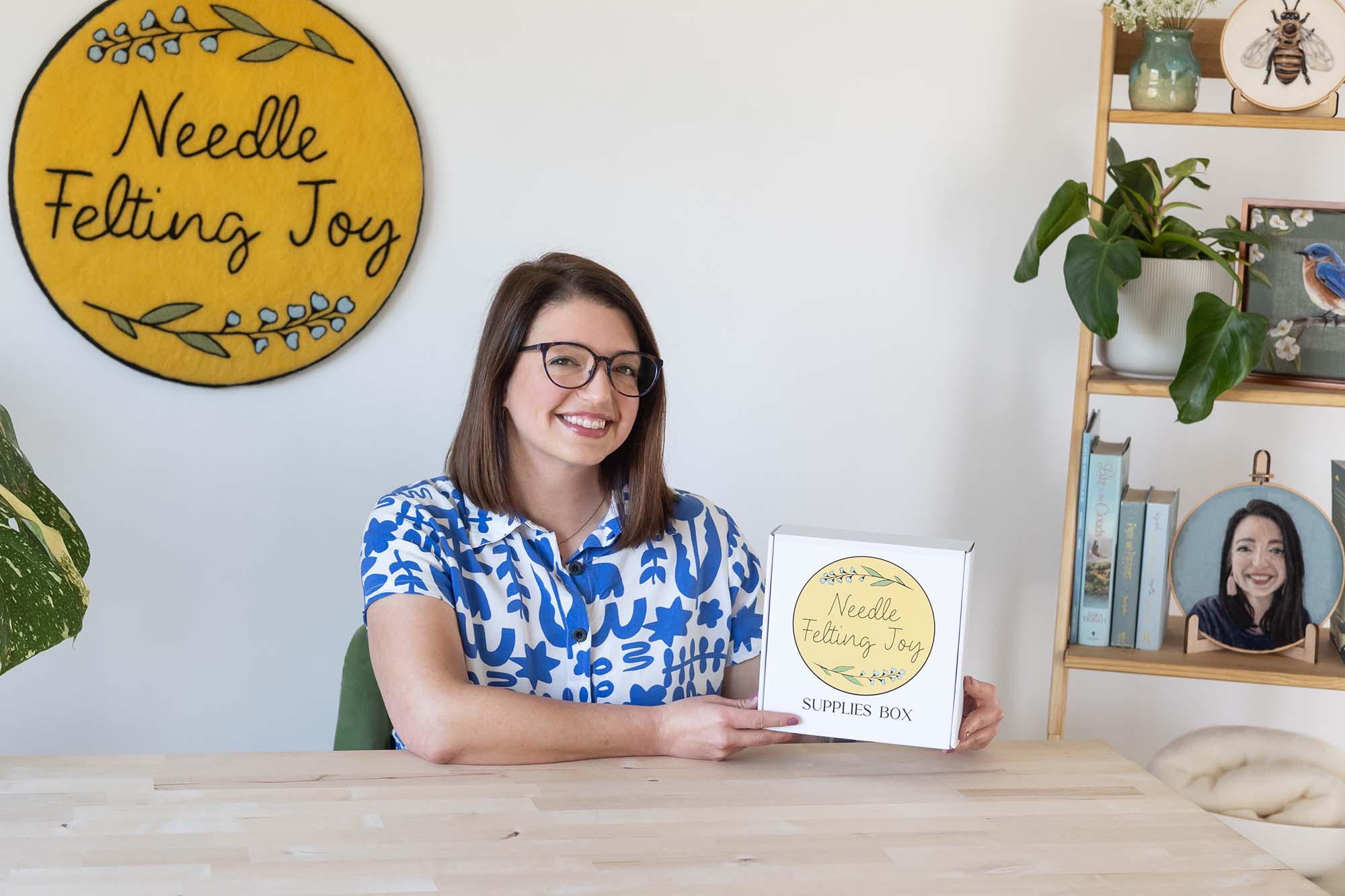 Woman holding a box titled 'Needle Felting Joy' in a room with decorative elements.
