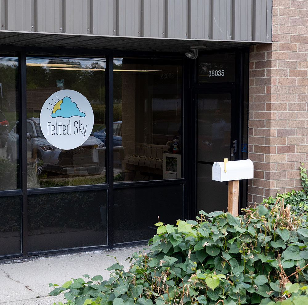 Storefront with 'Felted Sky' logo on glass door, surrounded by greenery.